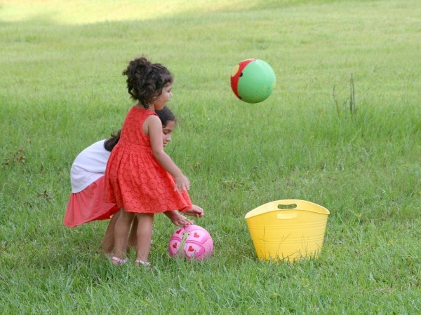Backyard-ball-games-for-kids - The Educators' Spin On It two girls tossing balls into yellow bucket in backyard